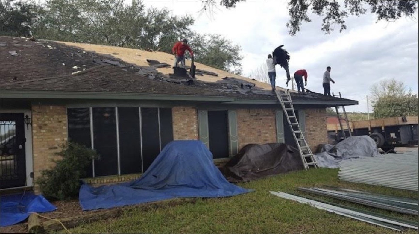 Crew removing old shingles during a roof tear-off, with new roofing panels staged on the ground